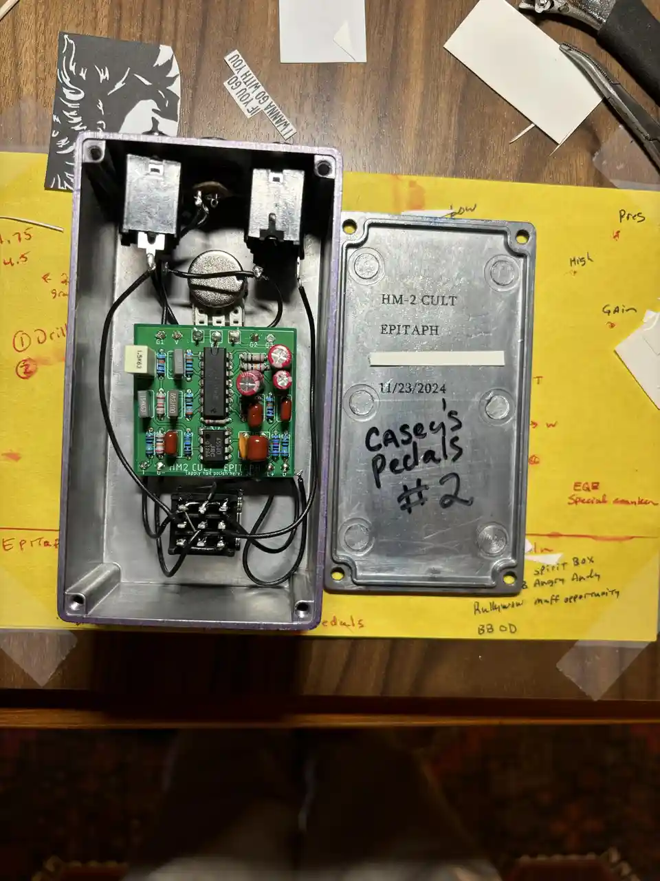 An open, hand-assembled guitar effects pedal with a green circuit board and silver metal casing sits on a yellow work…