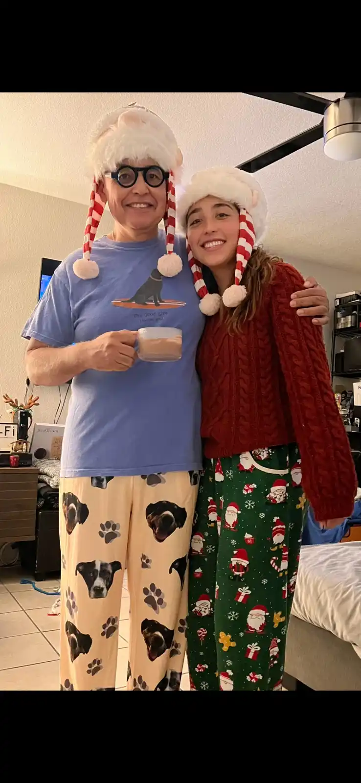 Two people are posing indoors wearing matching festive hats with red-and-white striped ear flaps and white pom-poms. The…