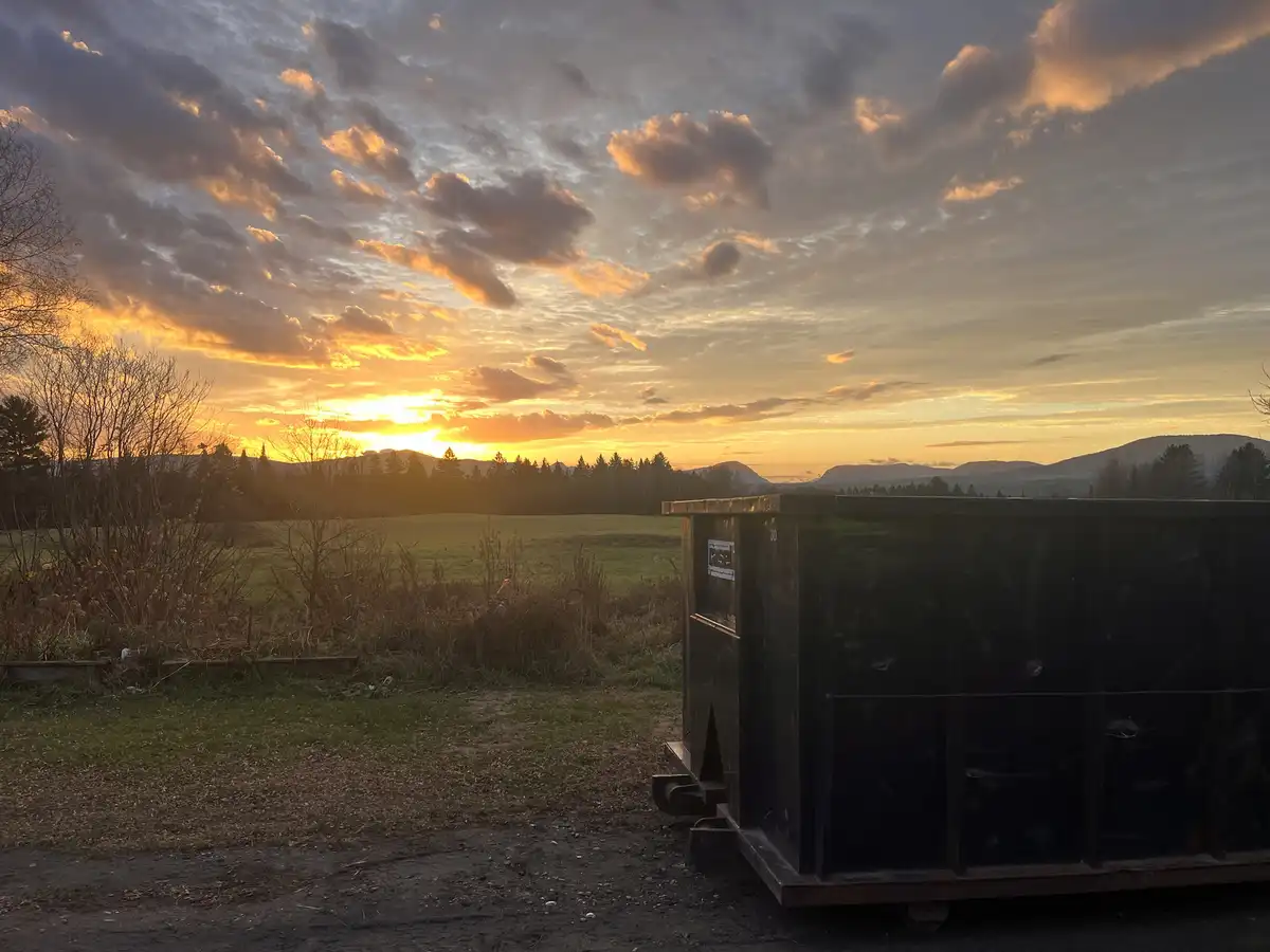 A large black dumpster sits in the foreground on a dirt path, blocking the view of a stunning sunset over a grassy field and…
