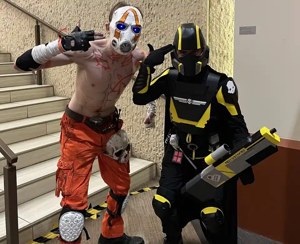 Two people in cosplay pose indoors near stairs; one wears an orange jumpsuit with red body paint and a white mask with…