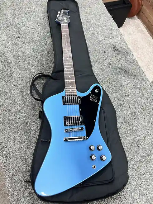 A light blue electric guitar with a black pickguard and four control knobs rests inside a black padded case on a gray…