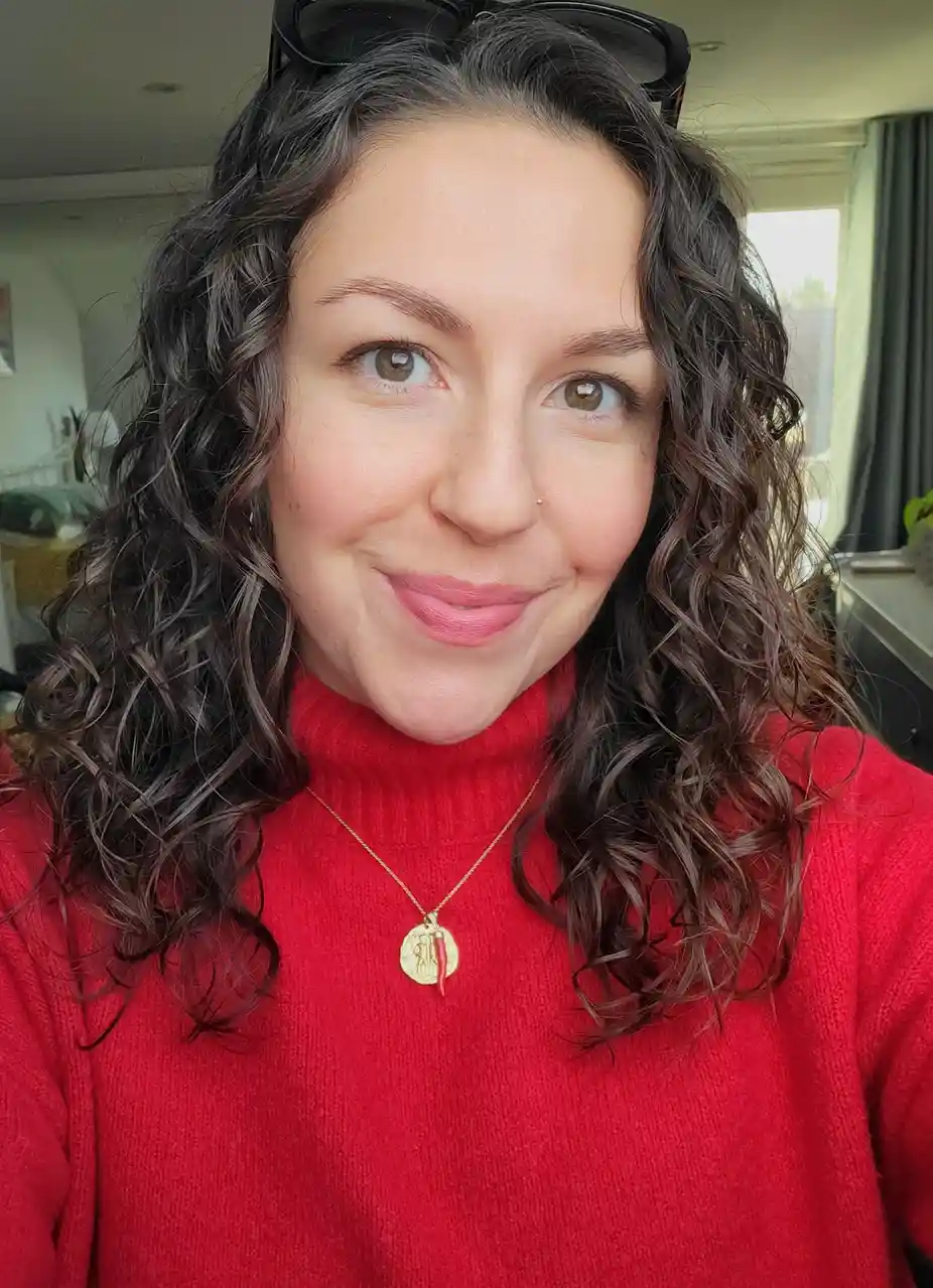A woman with dark curly hair and sunglasses on her head smiles at the camera while wearing a red turtleneck sweater. She has…