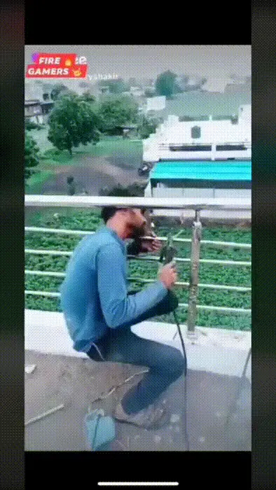 A man in a blue shirt is sitting on a balcony railing, holding a welding torch that emits bright sparks. He then leans…