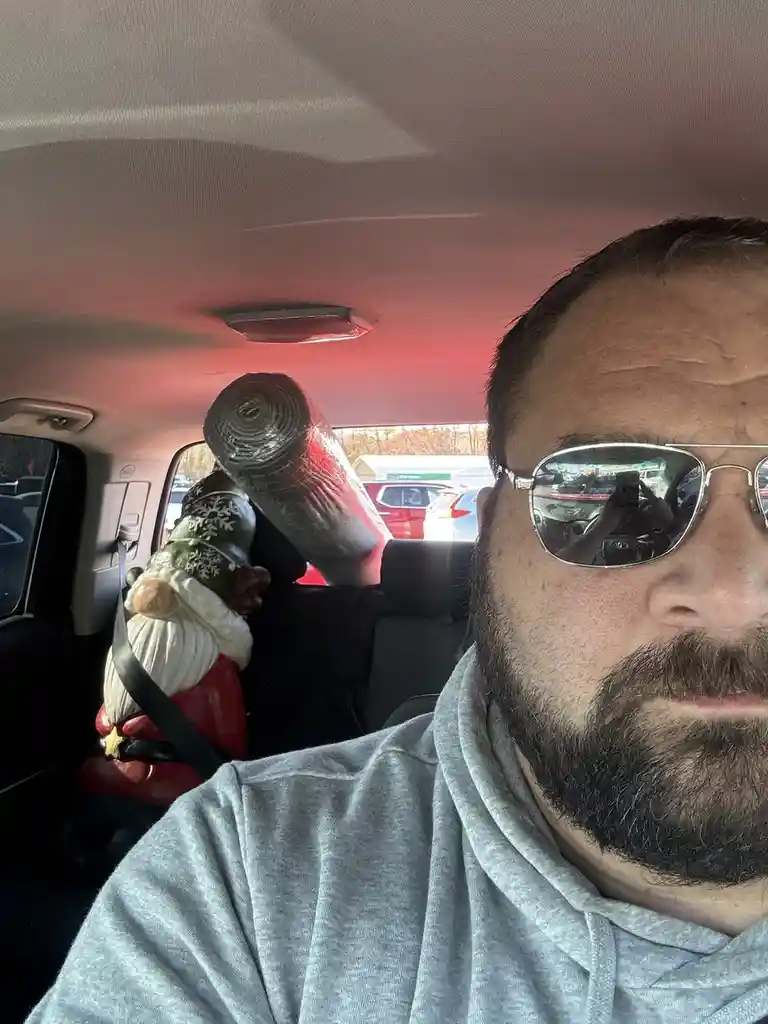 A man in a grey hoodie and sunglasses takes a selfie in a car, with a large rolled-up piece of insulation strapped to the…