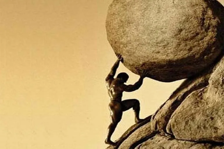 A bronze statue depicts a muscular man, Sisyphus, straining to push a massive boulder uphill on a rocky slope. The image is…