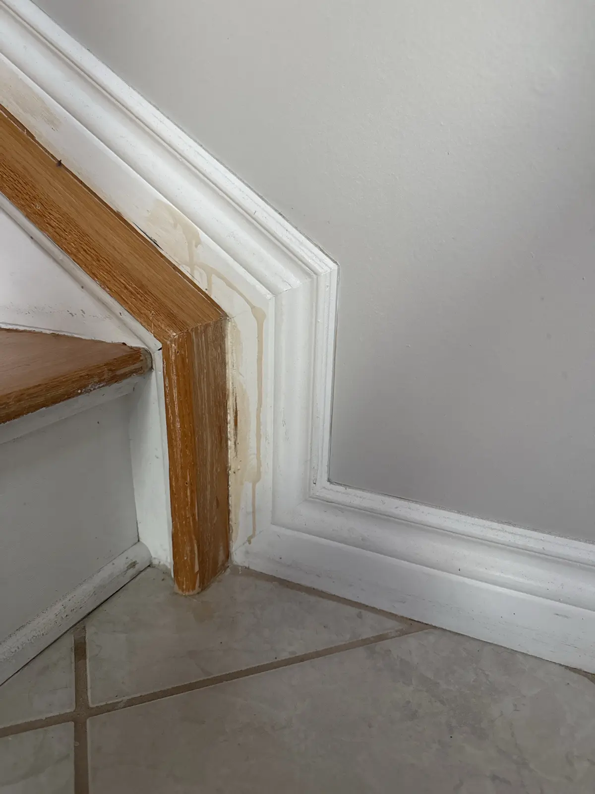A close-up photo shows water damage on a white-painted wooden staircase corner, with brownish stains dripping down the trim…