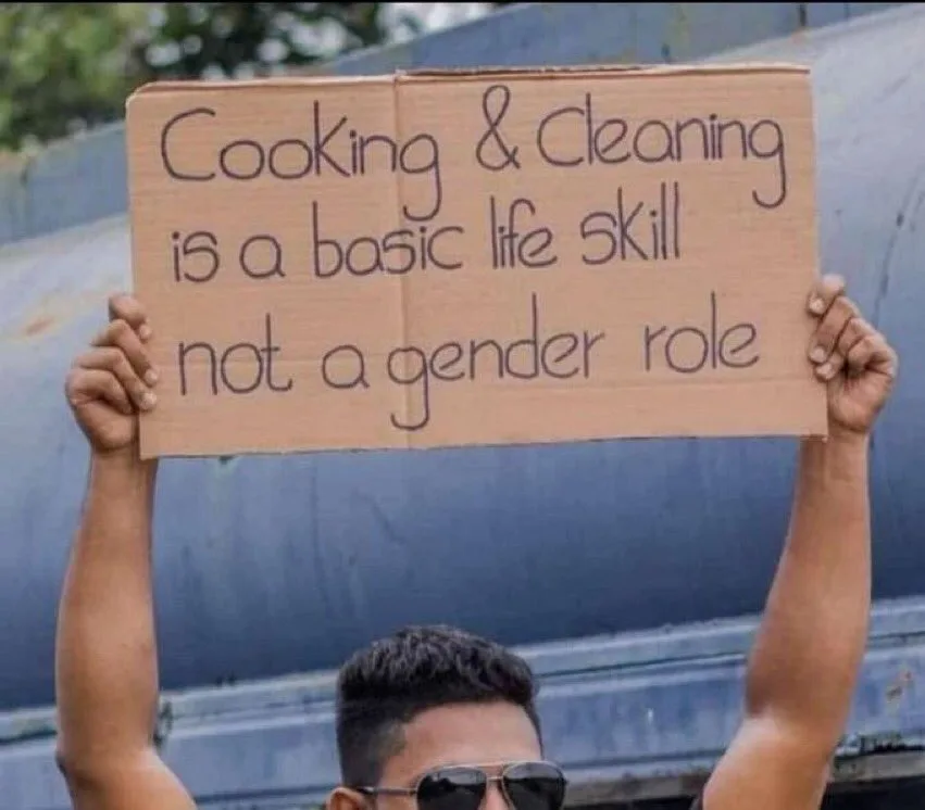 A person wearing sunglasses holds up a handmade cardboard sign with handwritten text that reads 'Cooking & Cleaning is a…