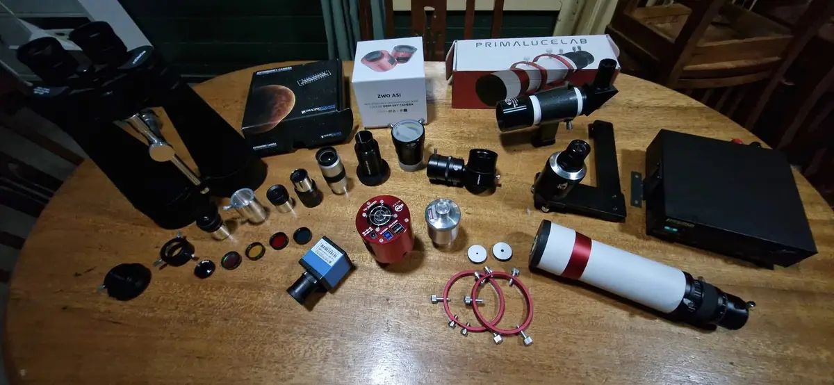 A wooden table is covered with an extensive collection of astronomy equipment, including telescopes, camera boxes labeled…