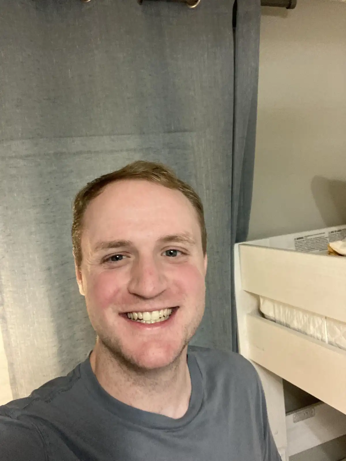 A man with reddish-brown hair and a wide, toothy grin takes a selfie in front of a grey curtain, with the headboard of a…