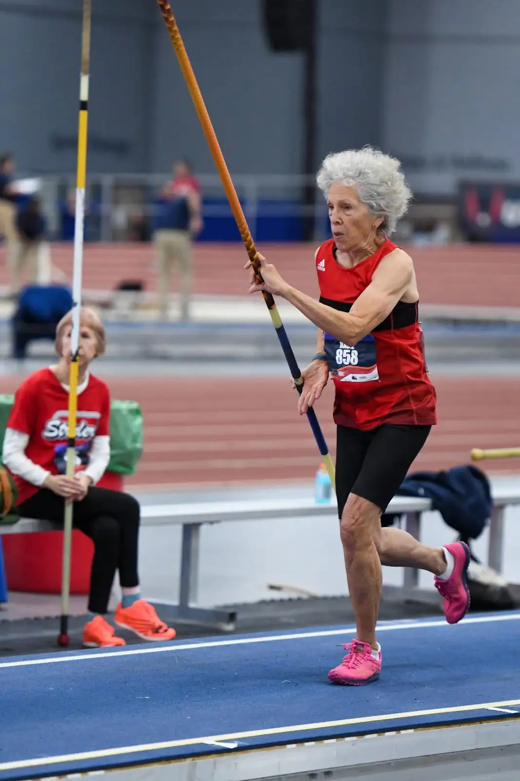 An elderly woman with white curly hair, wearing a red athletic tank top and black shorts, is mid-stride in a pole vaulting…