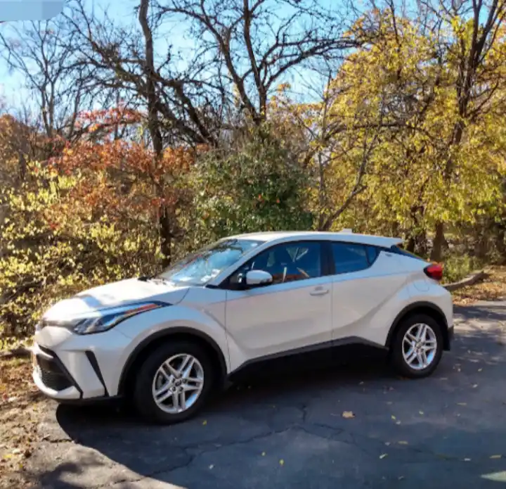 A white Toyota C-HR SUV is parked on a cracked asphalt road surrounded by autumn trees with yellow and orange leaves. The…