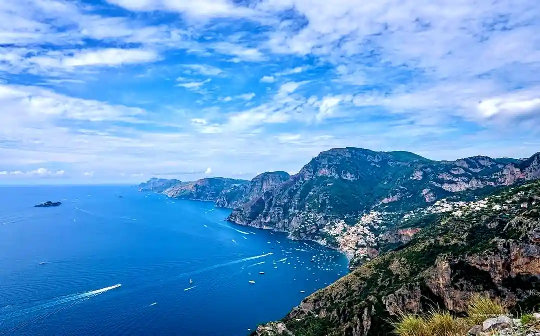 A scenic coastal view of a cliffside town with blue water, boats, and mountains under a partly cloudy sky. Overlaid text…