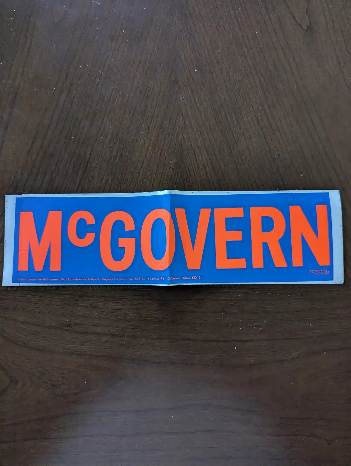 A vintage blue and orange political sticker for McGovern lies folded on a dark wooden surface. The large orange text…
