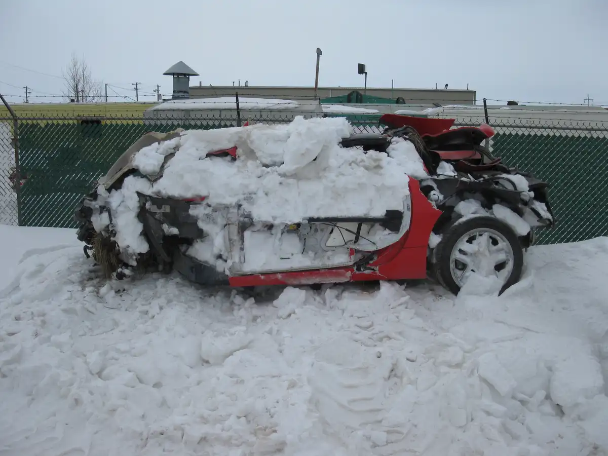 A red car is severely damaged and partially buried in snow, with its roof and body panels crushed and exposed. The vehicle…