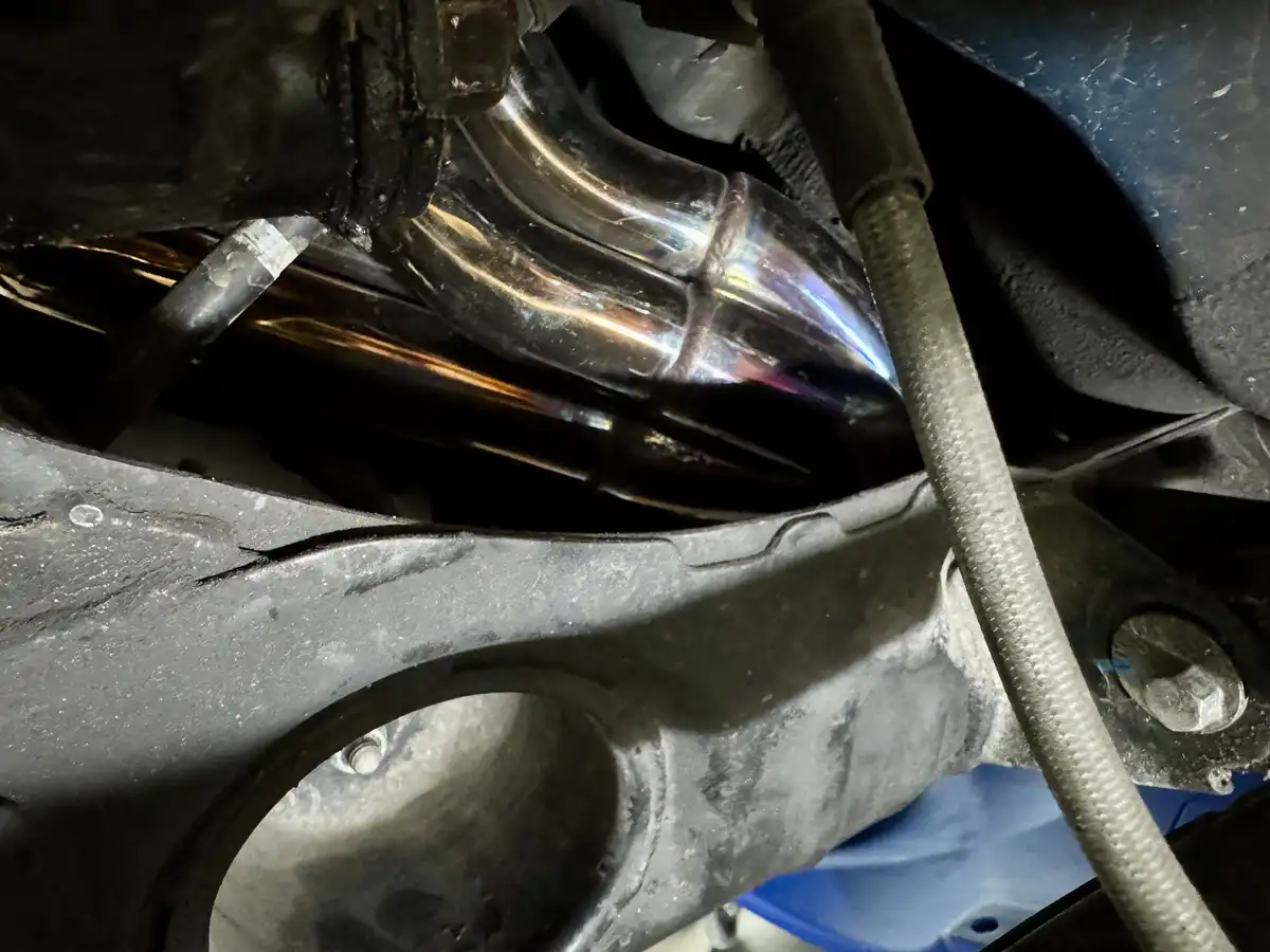 A close-up photo shows a shiny, heat-reflective exhaust pipe under a vehicle, glowing with iridescent colors from heat…