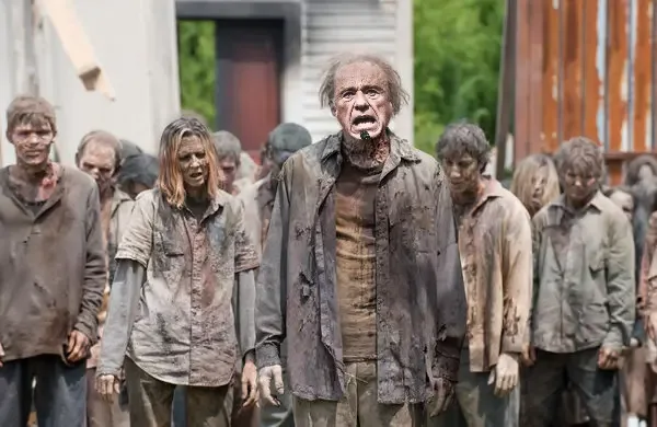 A man with a bloodied face and tattered clothes stands in the center of a crowd of dirty, disheveled zombies, looking upward…