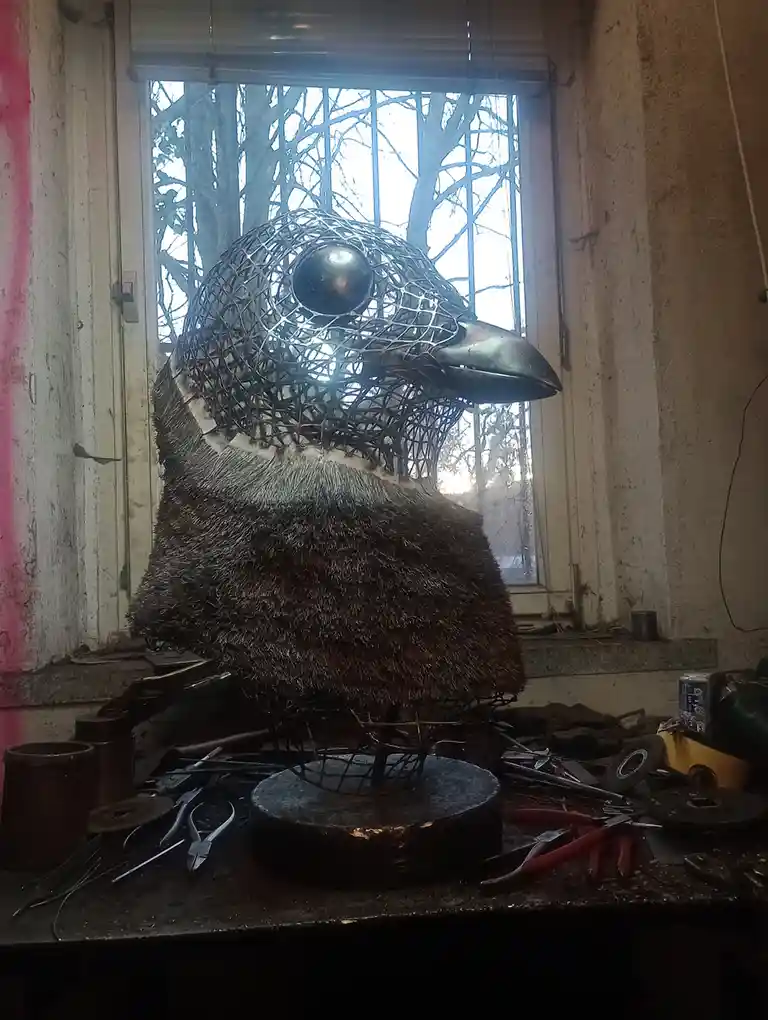 A partially constructed metal bird sculpture sits on a cluttered workbench in front of a window, its head made of wire mesh…