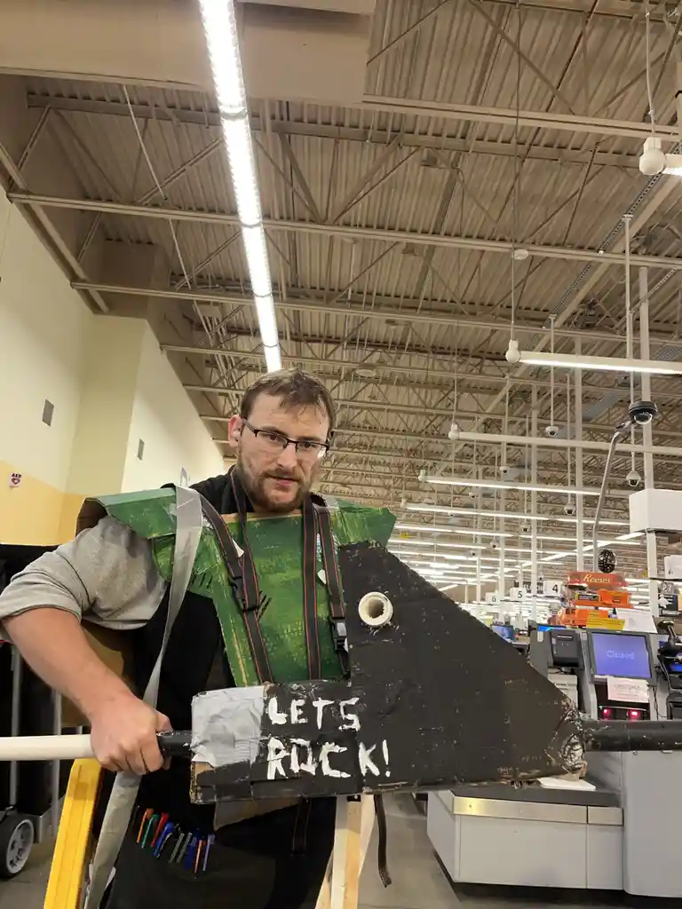 A man with glasses and a beard stands in a store aisle, wearing a homemade green and black cardboard costume resembling a…