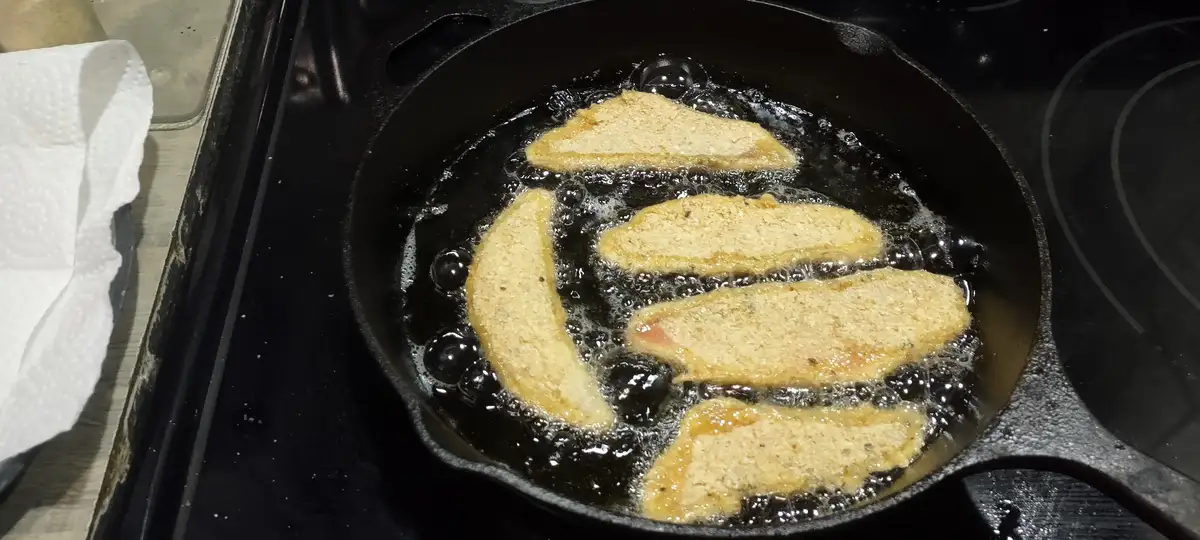 A black cast iron skillet sits on a stovetop, filled with hot oil where four breaded chicken tenders are frying. One tender…