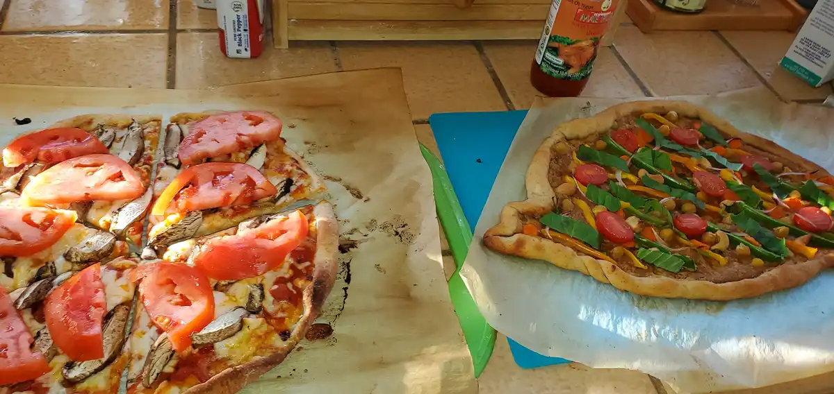 Two homemade pizzas sit side-by-side on parchment paper on a tiled kitchen counter. The pizza on the left is topped with…