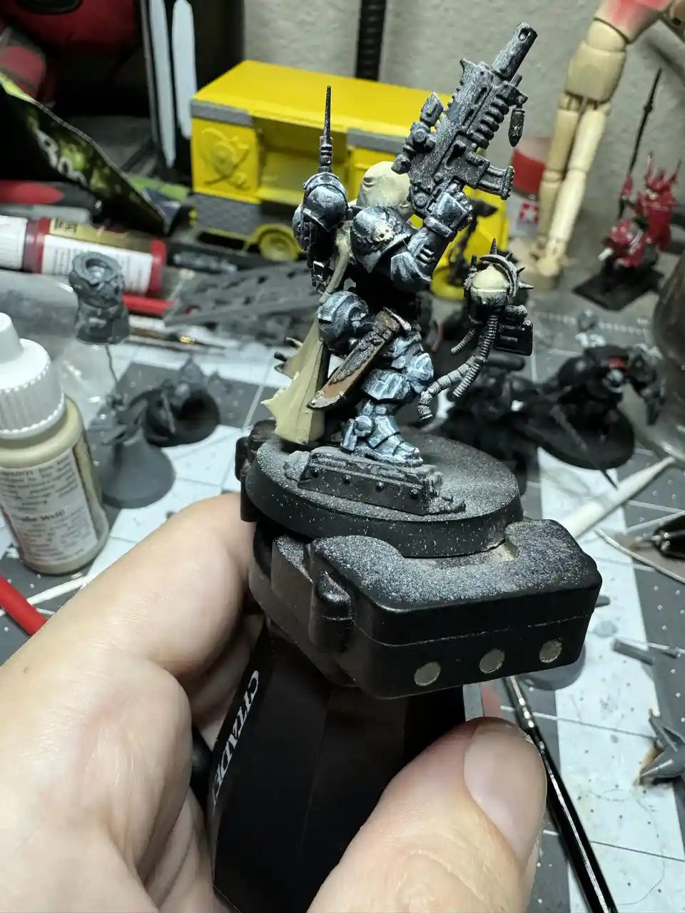 A hand holds a heavily weathered, unpainted miniature warhammer figurine mounted on a black base, surrounded by hobby tools…
