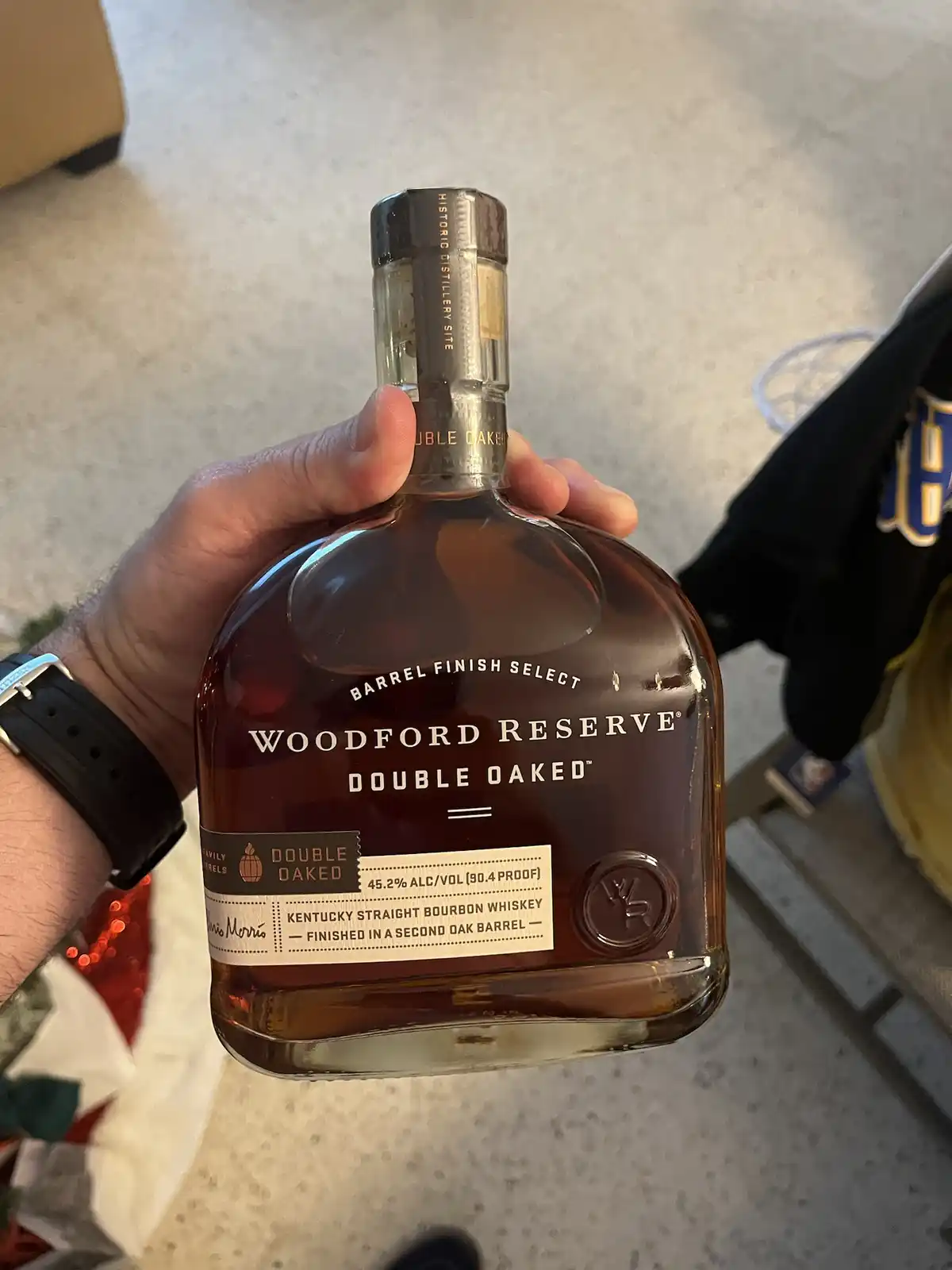 A hand holds a bottle of Woodford Reserve Double Oaked bourbon whiskey, showing its label and branding. The bottle is…