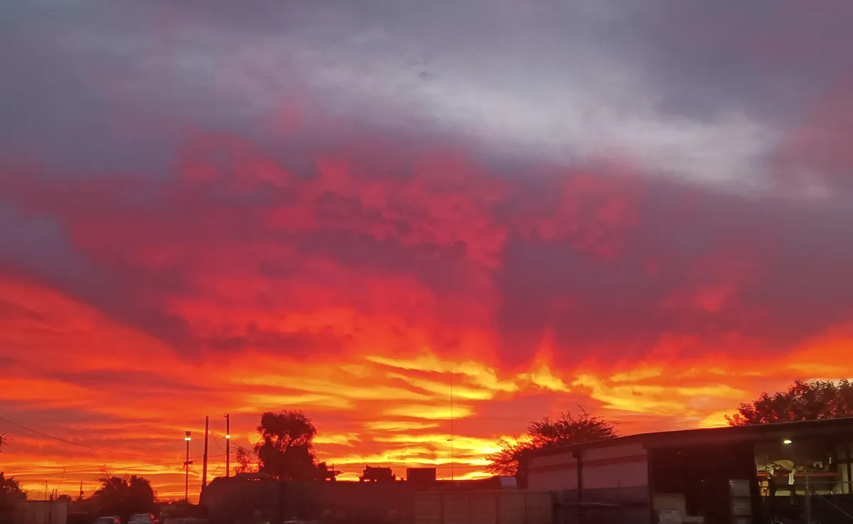 A dramatic, fiery sunset with clouds glowing in shades of red, orange, and purple over a silhouette of industrial buildings…