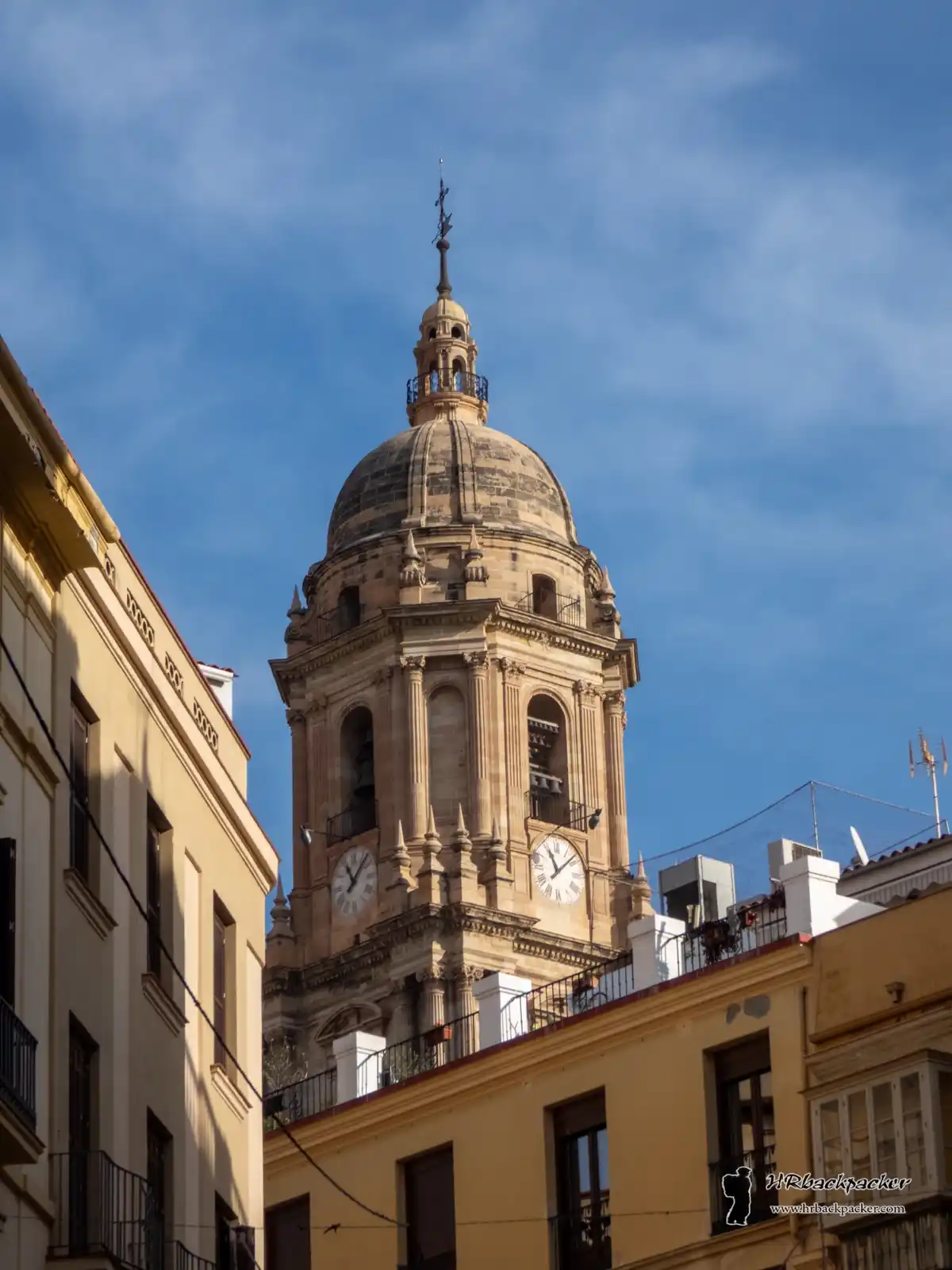 A photograph of a historic stone clock tower with a dome and spire, set against a blue sky, flanked by yellow buildings. The…