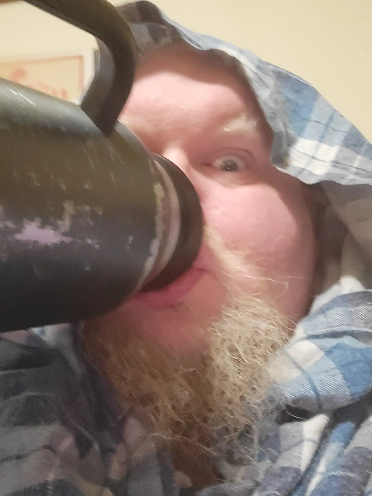 A man with a long, light-colored beard and a blue plaid shirt is shown drinking from a large black thermos. He is looking…