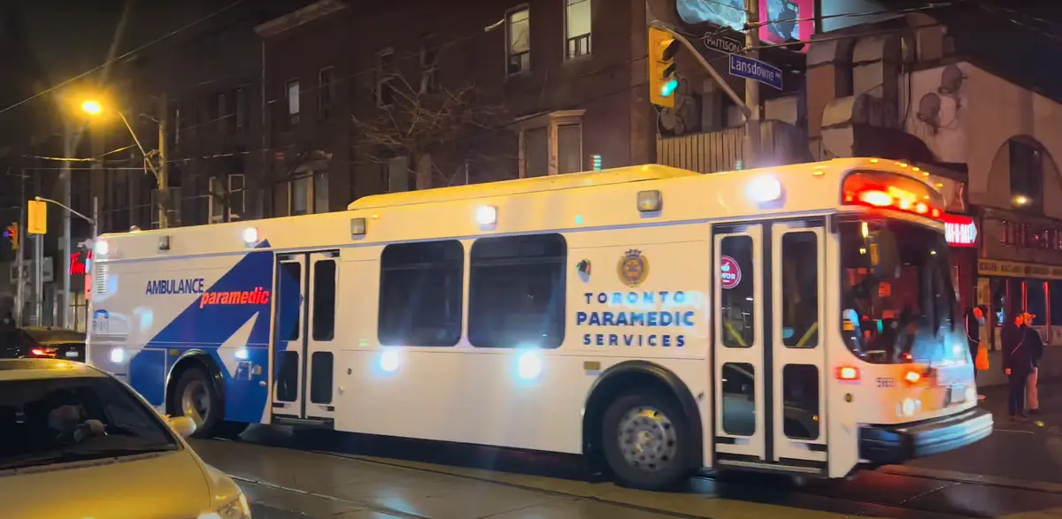 A white Toronto Paramedic Services ambulance bus, modified to look like a public transit bus, is shown at night on a city…