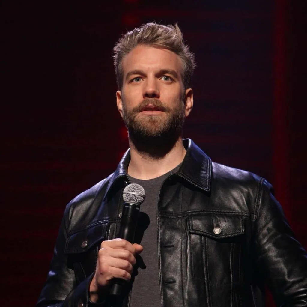 a man with a beard and short blond hair wearing a black leather jacket over a gray shirt holds a microphone on a stage with…