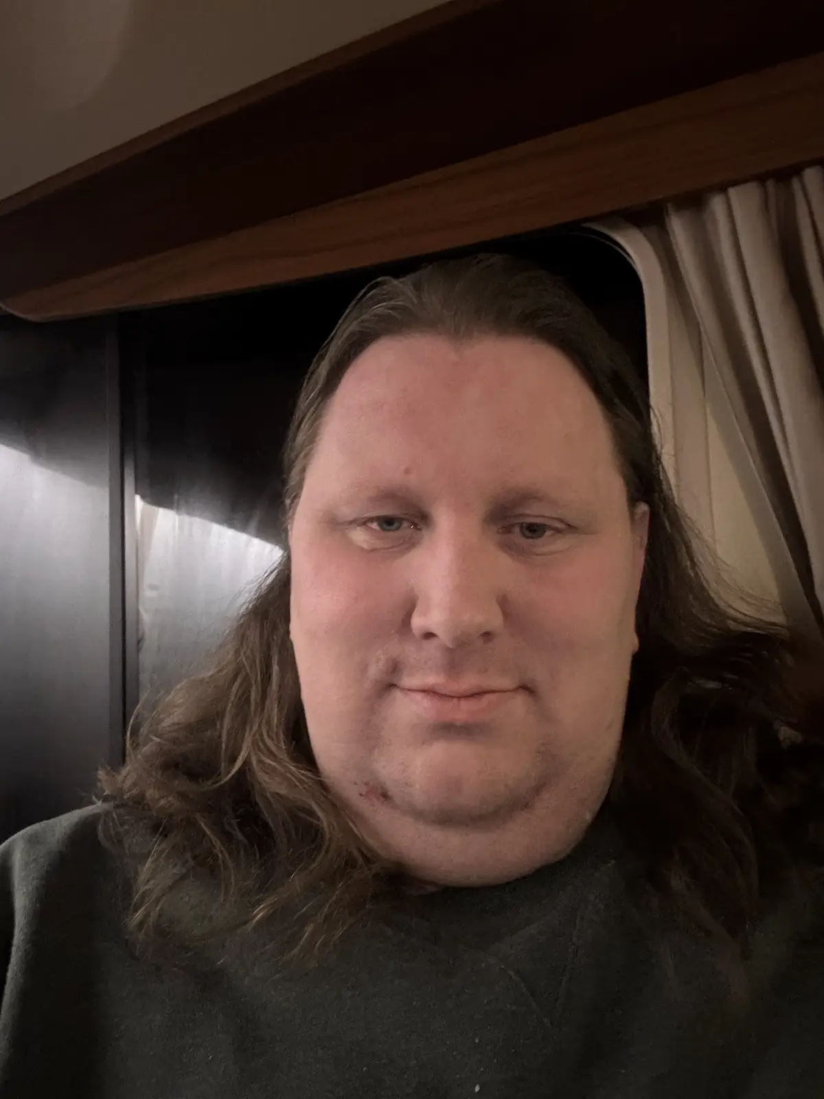 A close-up selfie of a man with long brown hair and a neutral expression, wearing a dark grey shirt, taken inside what…