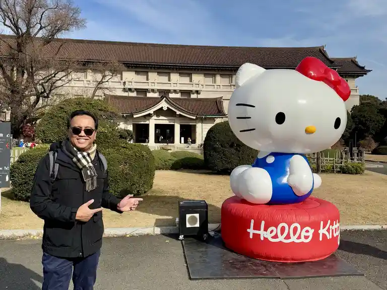 A man in a black jacket and scarf stands smiling and gesturing toward a giant inflatable Hello Kitty statue in front of a…