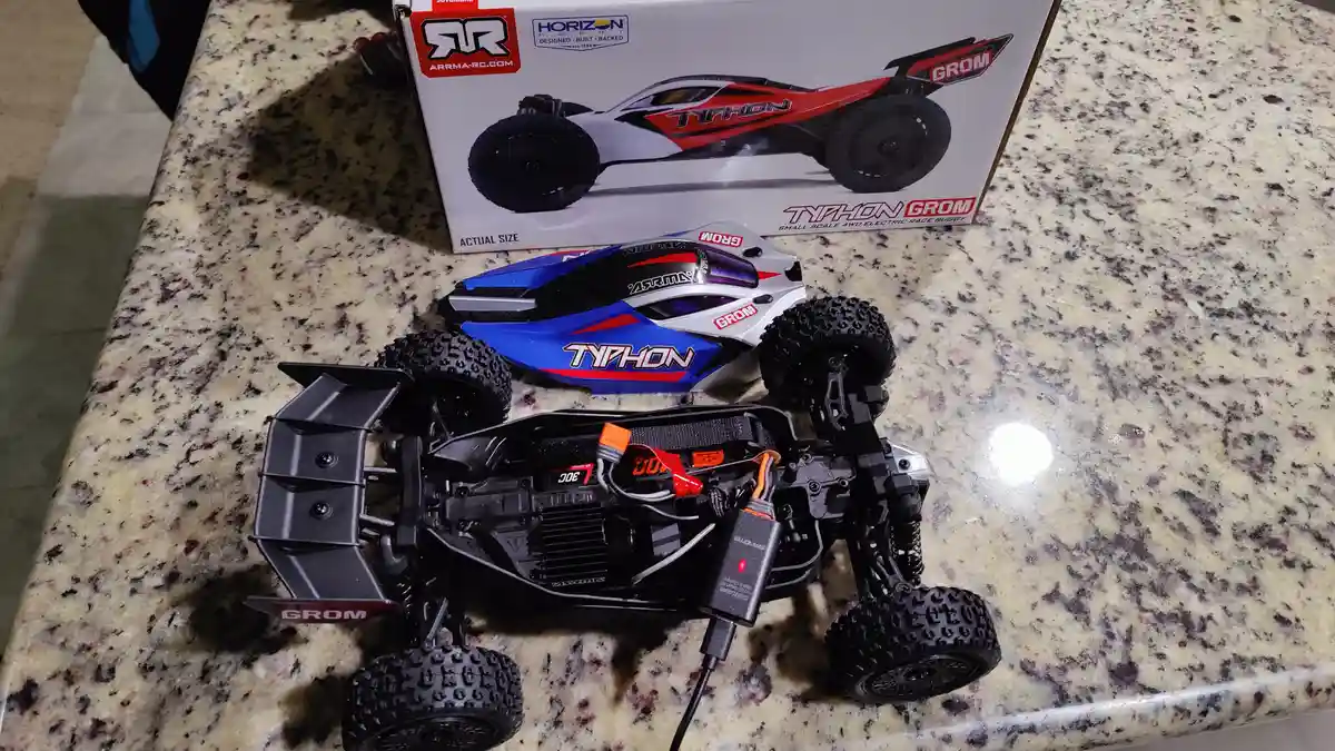 A disassembled blue, white, and red Typhon Grom remote-controlled buggy sits on a speckled countertop with its box behind…