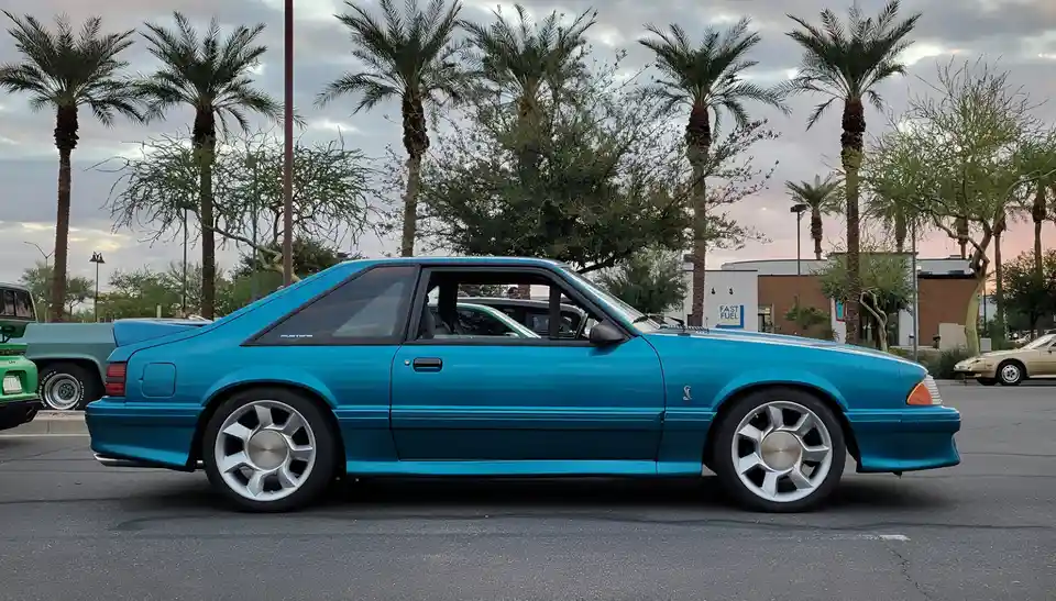 A bright blue 1990s Ford Mustang SVO is parked in a lot with palm trees and other cars visible. The overlaid text reads 'i'm…