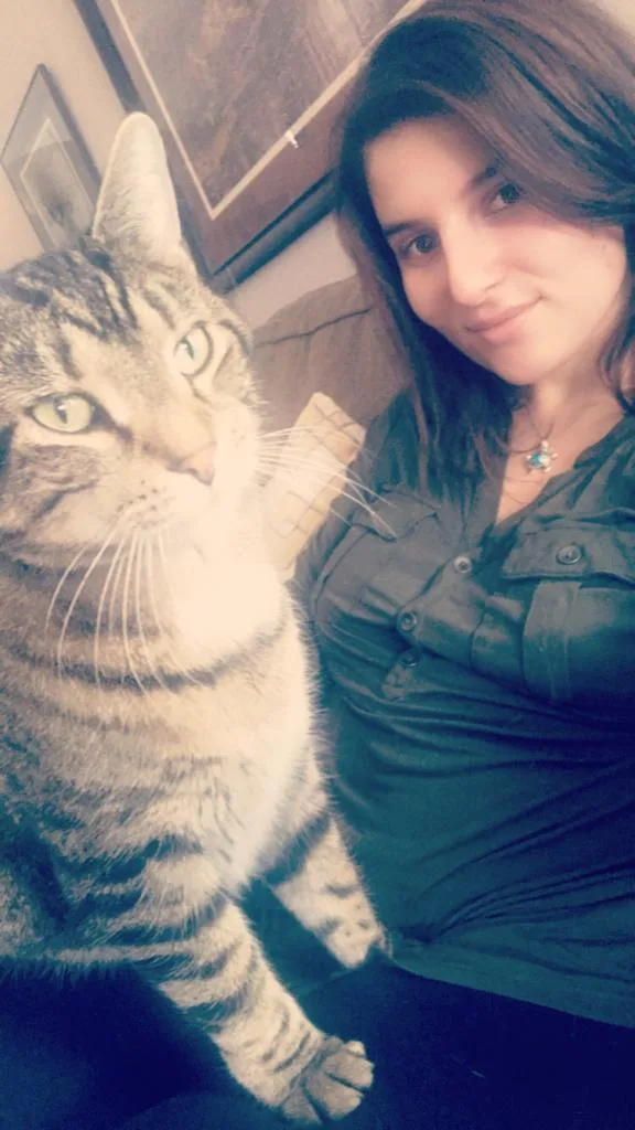A selfie shows a woman with dark hair and a green shirt smiling at the camera while holding a large tabby cat. The cat, with…