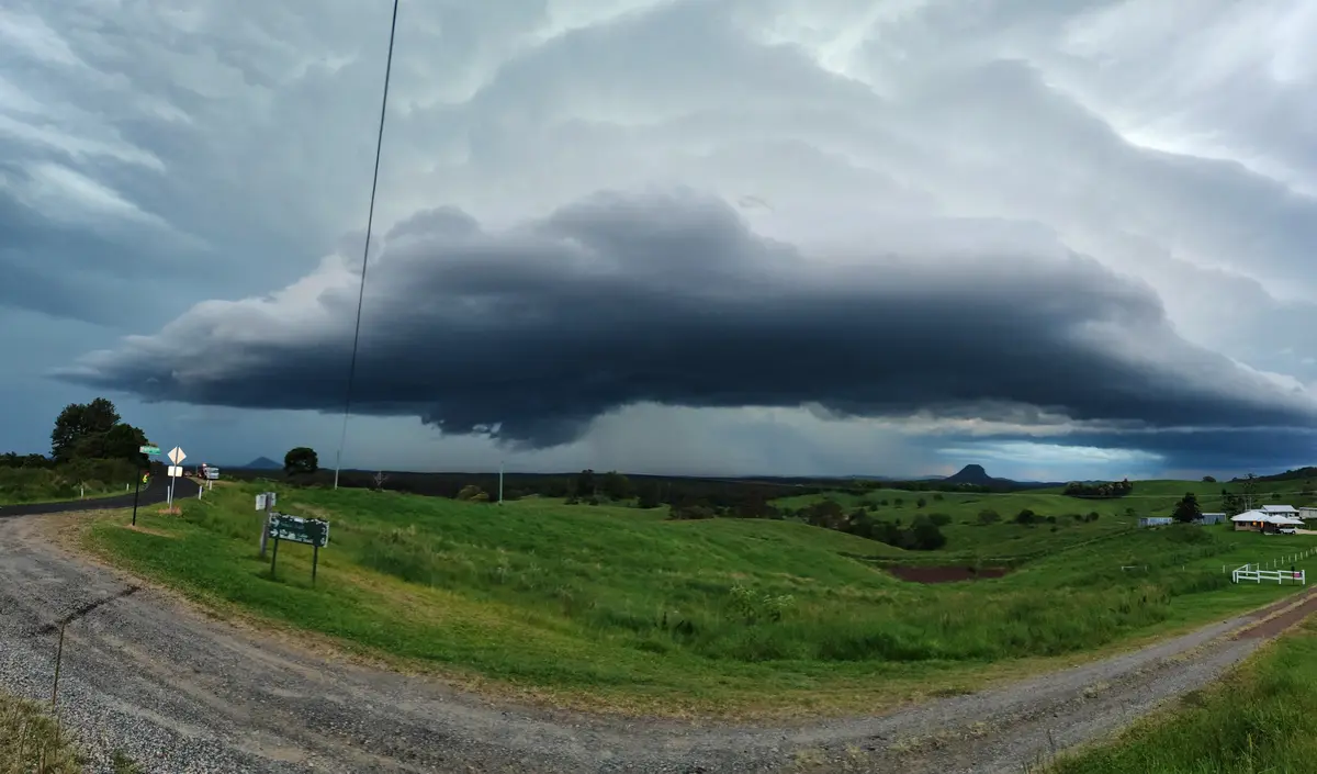 A wide-angle photo shows a dramatic, dark storm cloud looming over a rural landscape with green hills, gravel roads, and…