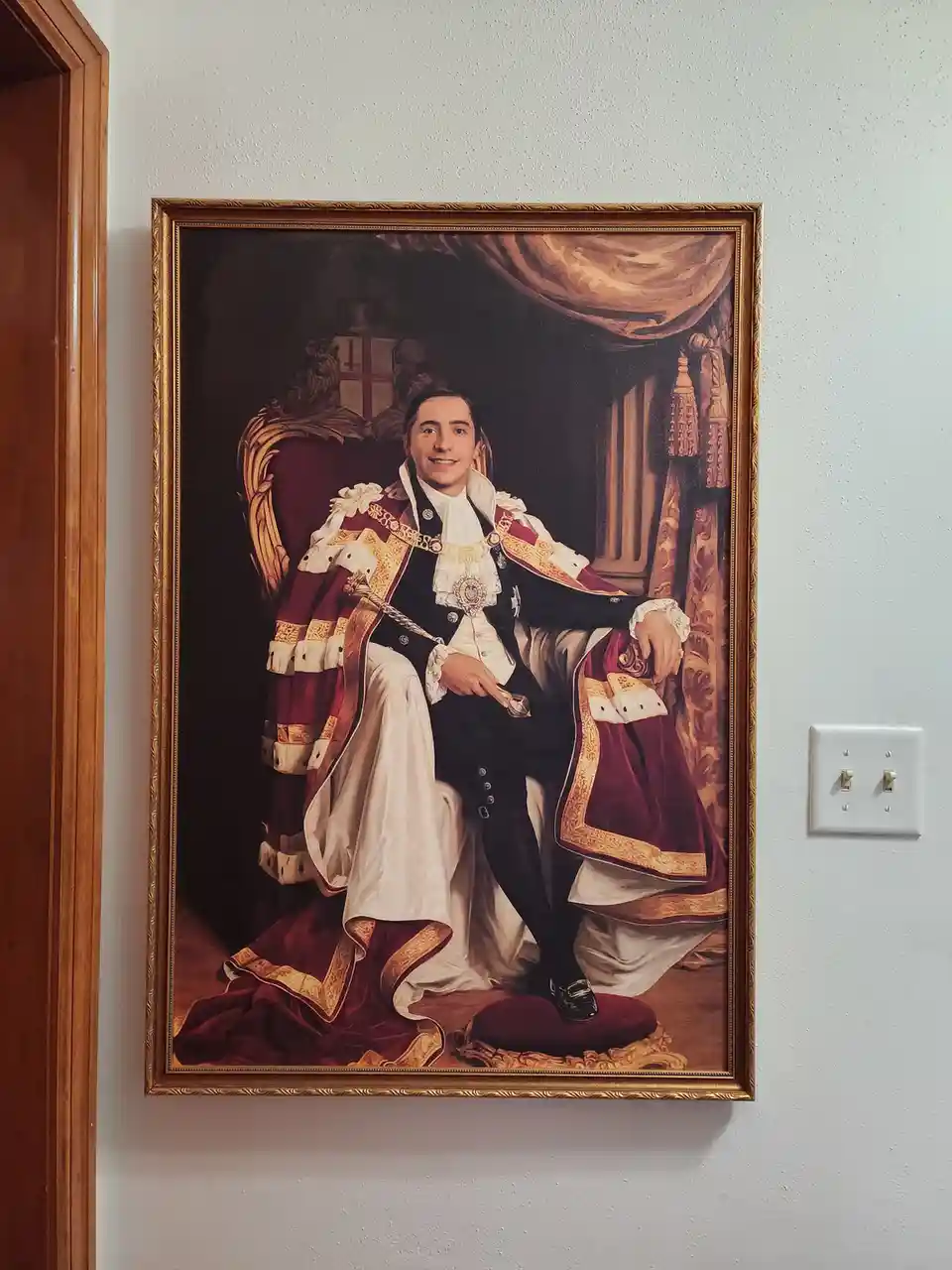 A framed portrait of a man resembling John Oliver dressed in elaborate royal regalia, seated on a throne, hangs on a plain…