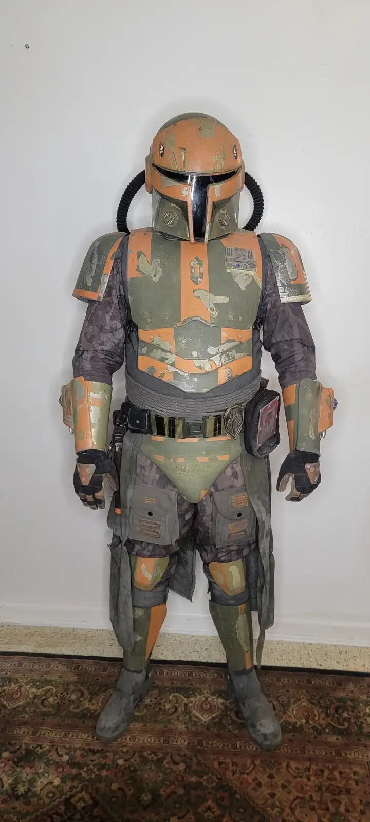 A full-body Boba Fett costume stands against a plain white wall on a patterned rug, its armor heavily weathered and painted…