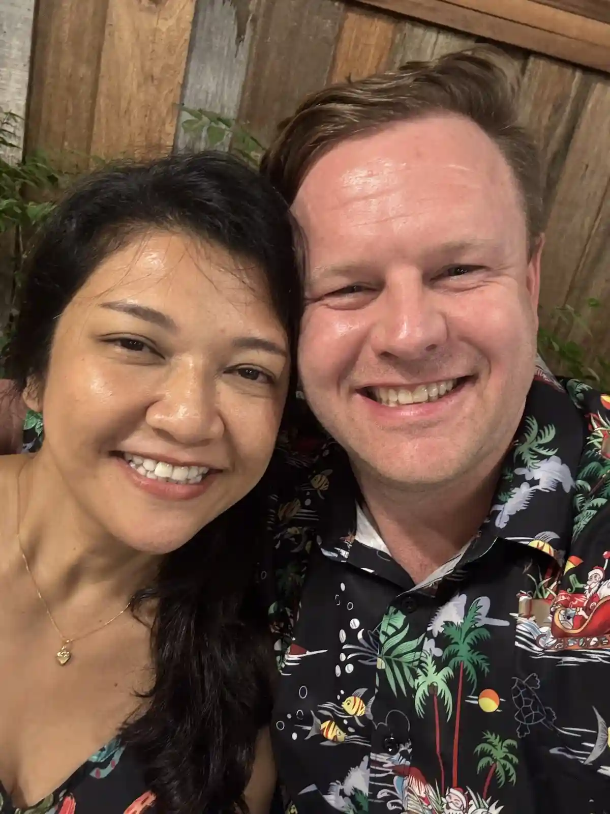 A selfie of a smiling man and woman with a wooden wall and greenery behind them. The man wears a black Hawaiian shirt with a…