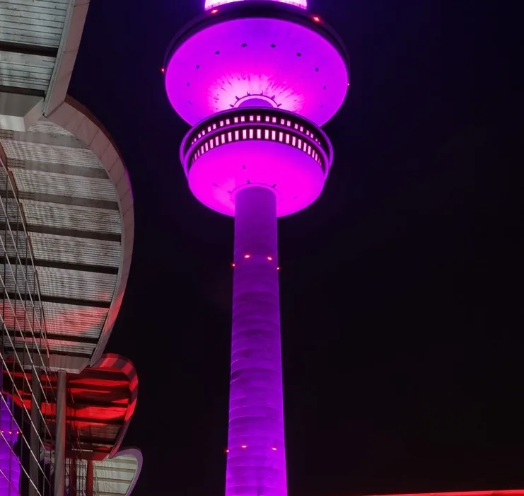 A tall, cylindrical tower illuminated in vibrant purple light against a dark night sky, with a modern building partially…