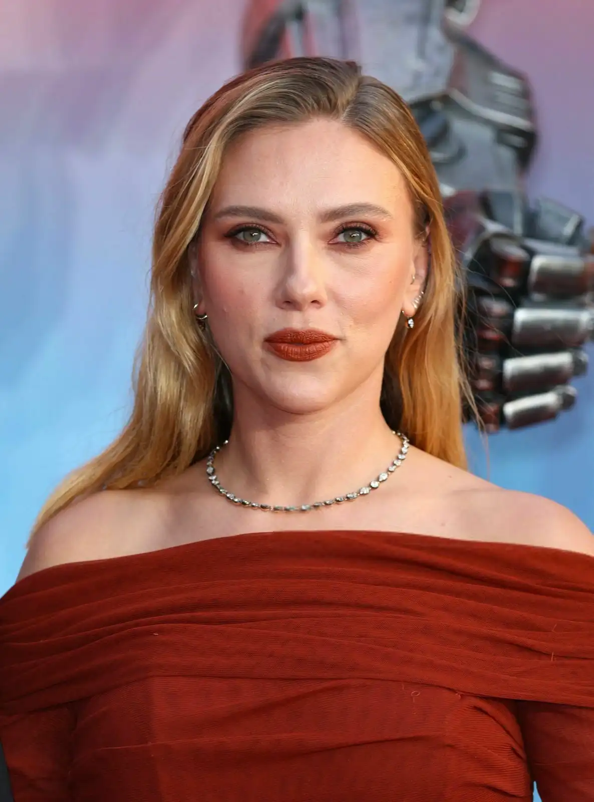 A woman with blonde hair and green eyes, wearing a rust-colored off-the-shoulder dress and a diamond necklace, looks…