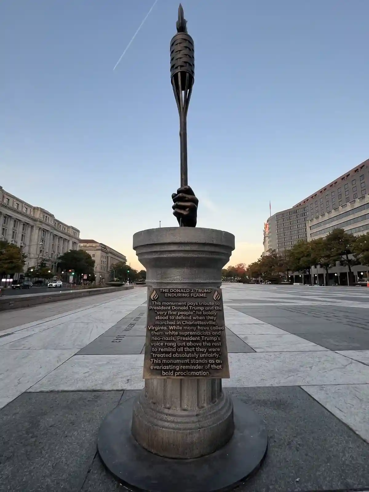 A photograph of a bronze monument featuring a hand gripping a torch, set on a stone pedestal in an urban plaza. The pedestal…