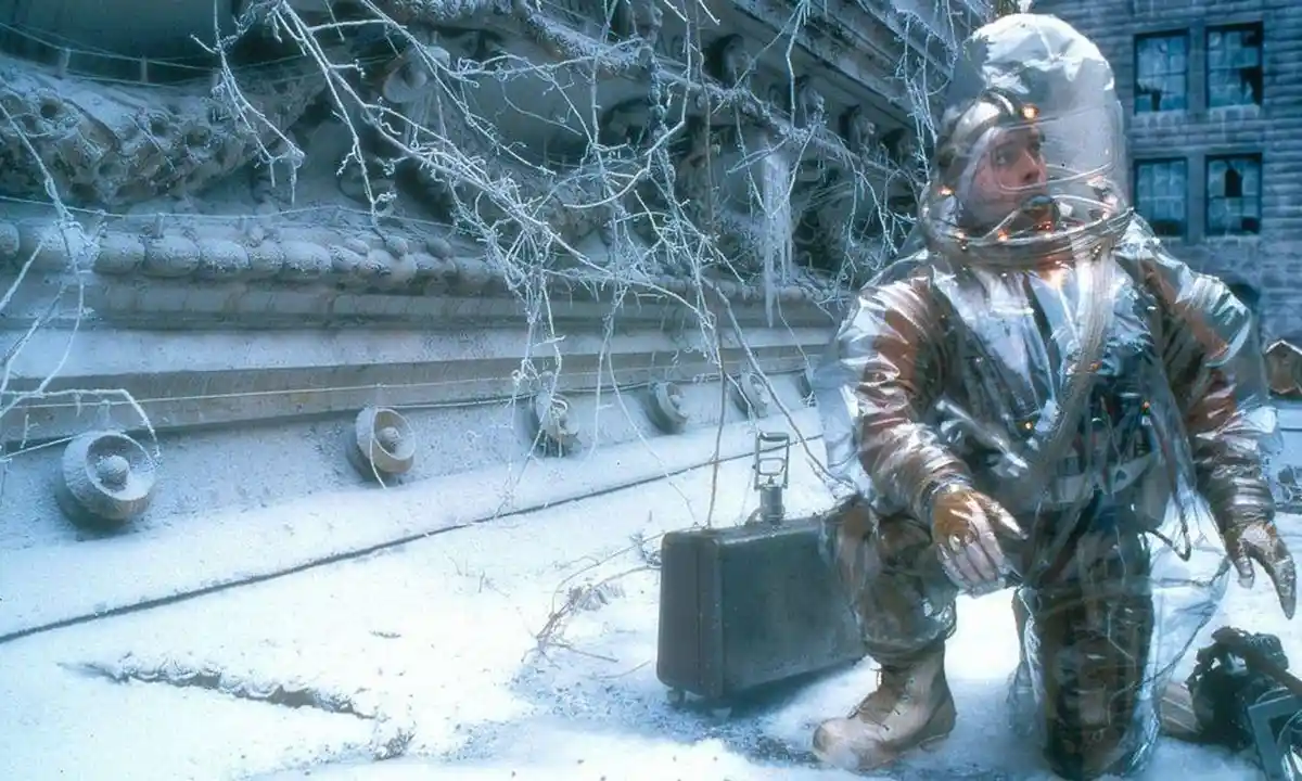 A man in a transparent, bulky space suit kneels in a snow-covered urban environment, next to a briefcase. Ice-covered…