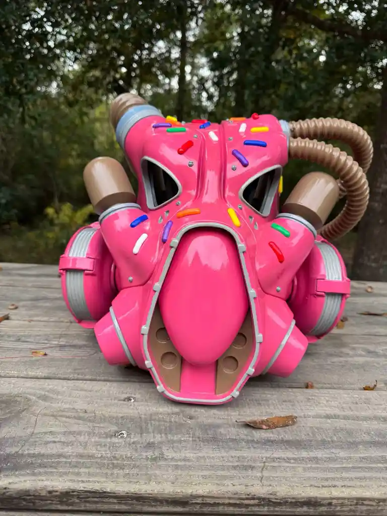 A bright pink, donut-shaped respirator mask with colorful sprinkles and brown tubes sits on a wooden table outdoors. The…