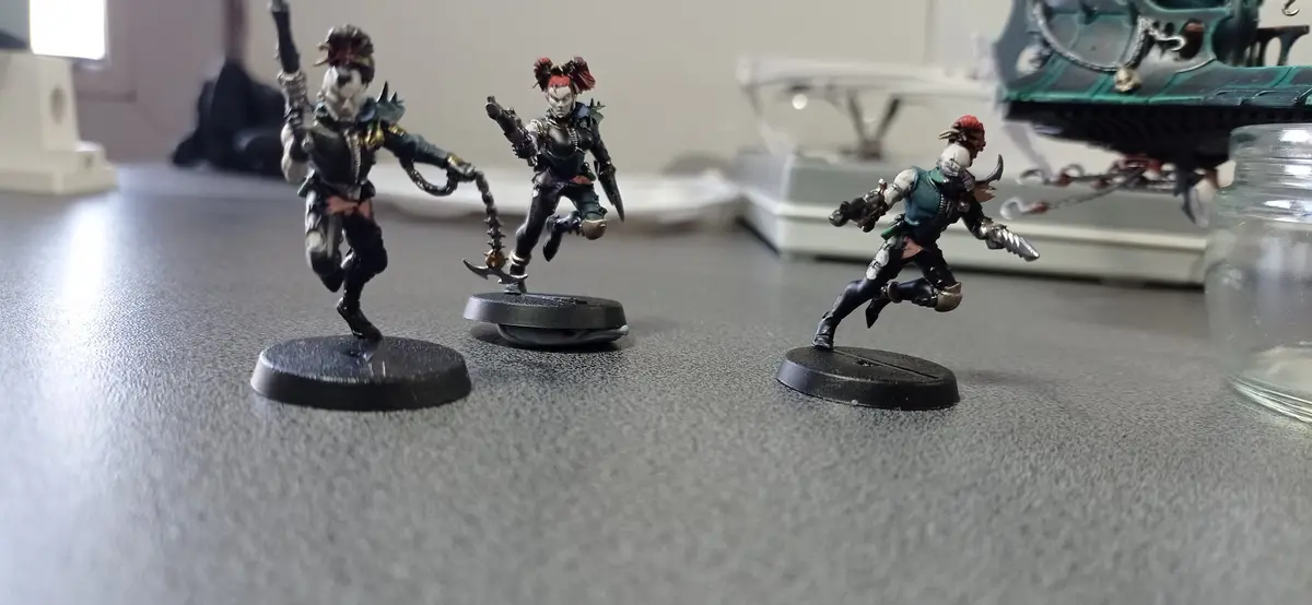 Three painted miniature figurines of fantasy or sci-fi characters are arranged in a line on a gray surface, each in a…