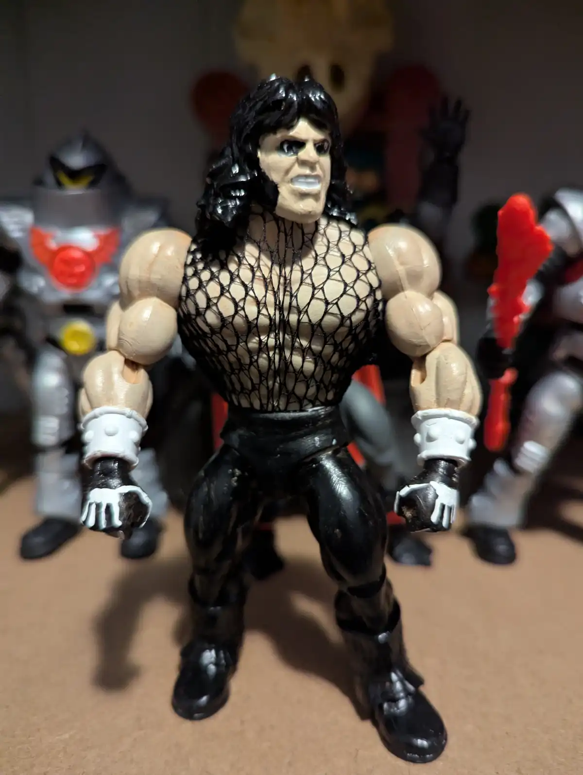 A muscular action figure with long black hair, wearing a black net top and black pants, stands in front of other action…