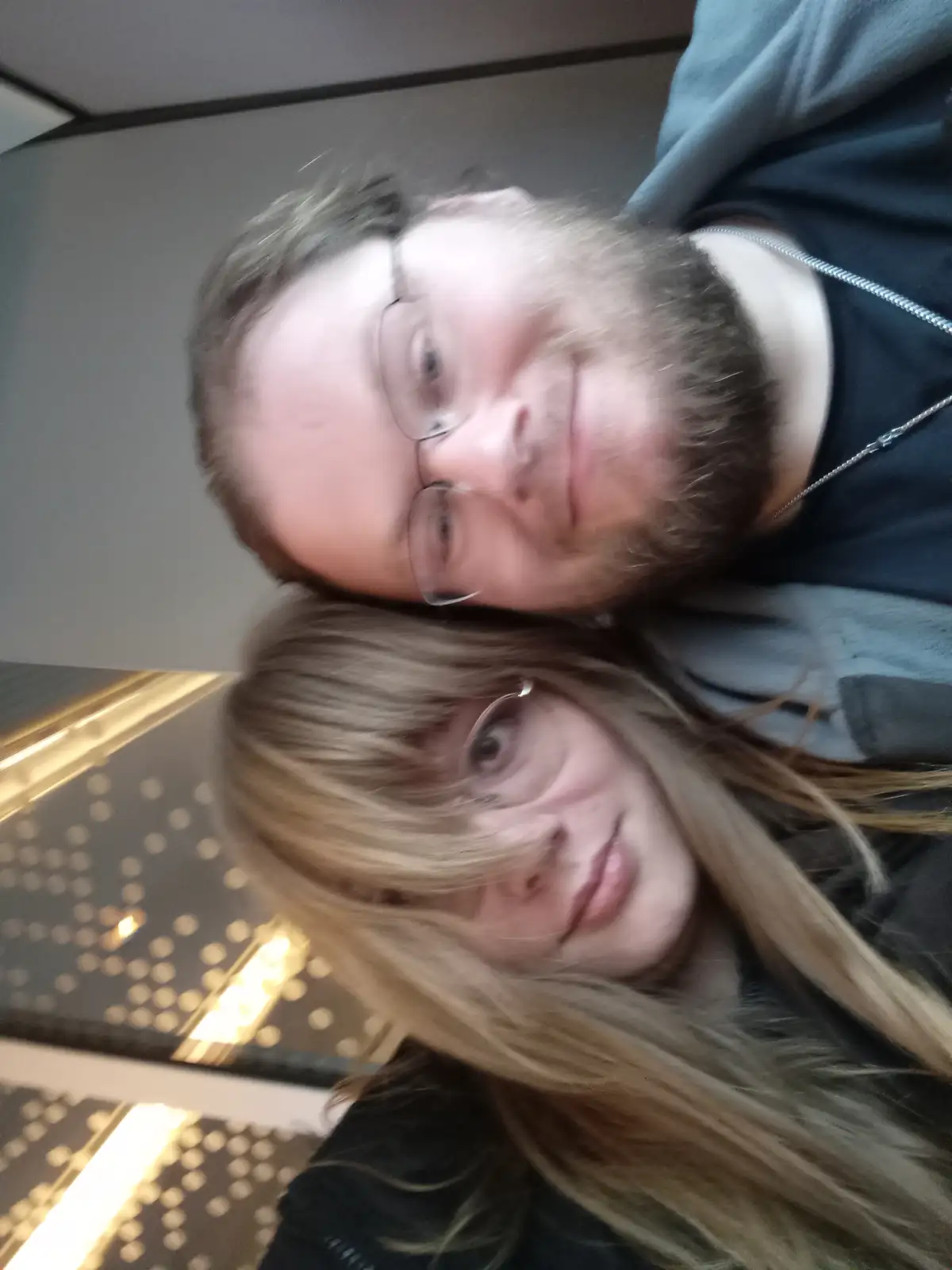 A blurry, tilted selfie shows a man with glasses and a beard lying above a woman with long blonde hair and glasses, both…