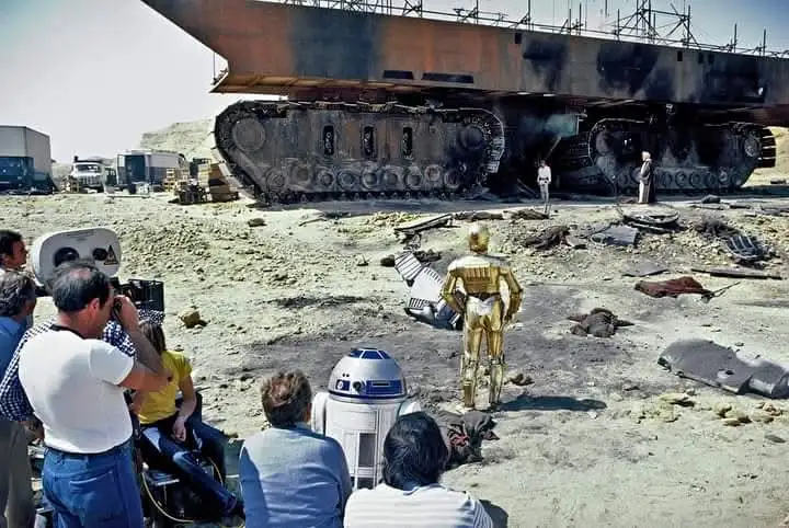 A behind-the-scenes photo from a Star Wars film set shows the golden droid C-3PO and the blue-and-white droid R2-D2 standing…