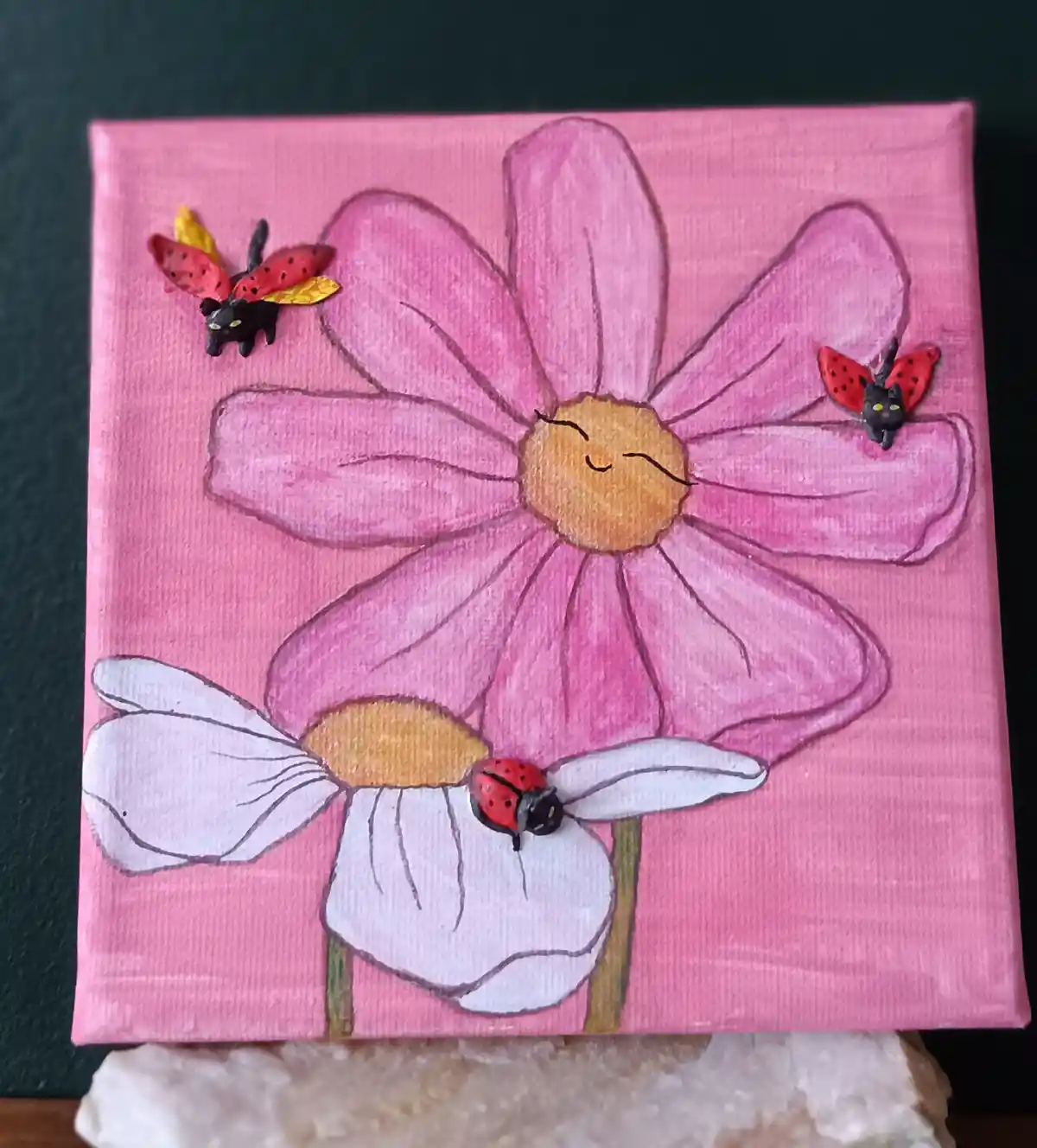 A hand-painted canvas depicts a large pink flower with a smiling face and a smaller white flower below. Three small…